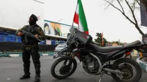 رجل مسلح في طهران، يرتدي زيا عسكريا وقناع وجه أسود، يحمل بندقية حربية. بجانبه دراجة نارية وخلفه صورة يظهر فيها وجه المرشد السابق للجمهورية علي خامنئي