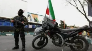 رجل مسلح في طهران، يرتدي زيا عسكريا وقناع وجه أسود، يحمل بندقية حربية. بجانبه دراجة نارية وخلفه صورة يظهر فيها وجه المرشد السابق للجمهورية علي خامنئي