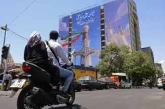 التعليق على الصورة،مارة في طهران قرب لافتة ضخمة تُظهر صواريخ إيرانية وسيف الإمام علي، وسط تصاعد التوتر بعد ضربات أمريكية جديدة على جنوب إيران.